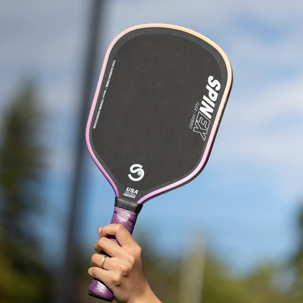 Player holding the Spinex FLEX Hybrid Storm pickleball paddle up against a blue sky on an outdoor court