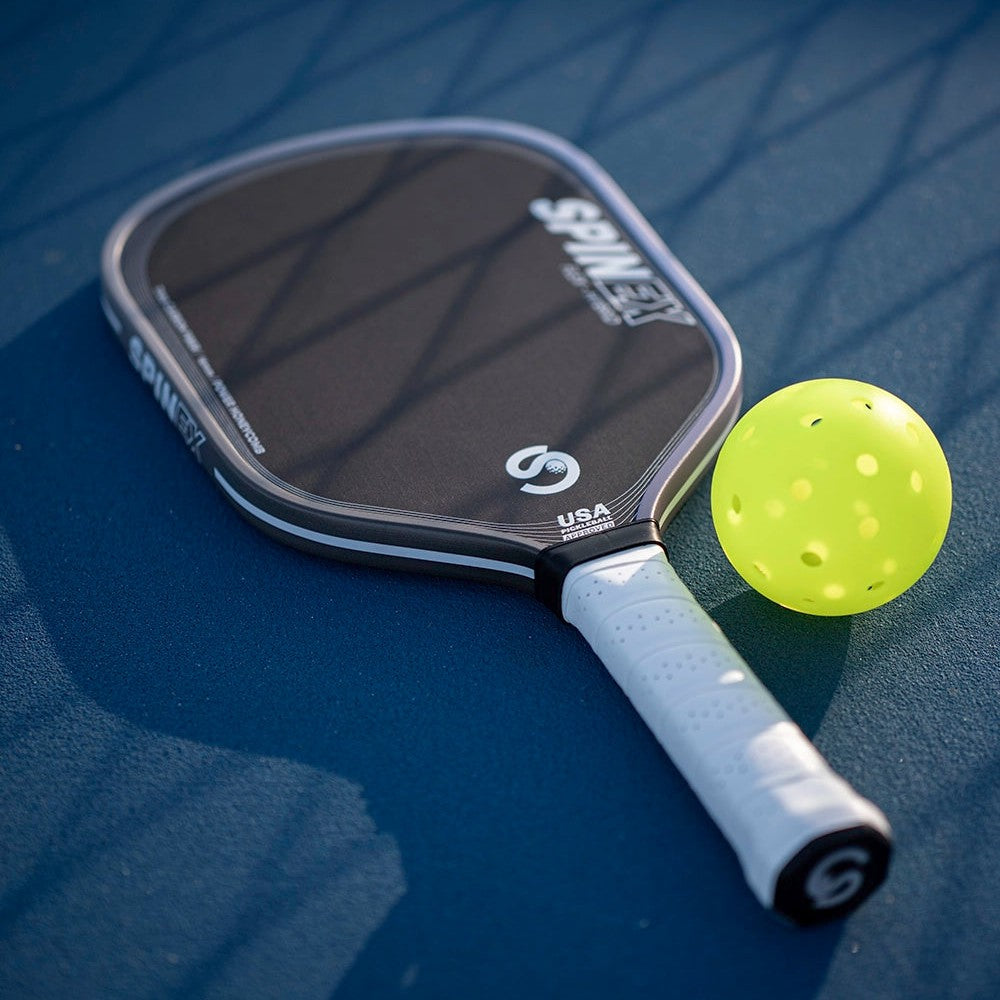Spinex FLEX Hybrid Iron pickleball paddle with white grip resting on a blue court beside a yellow pickleball